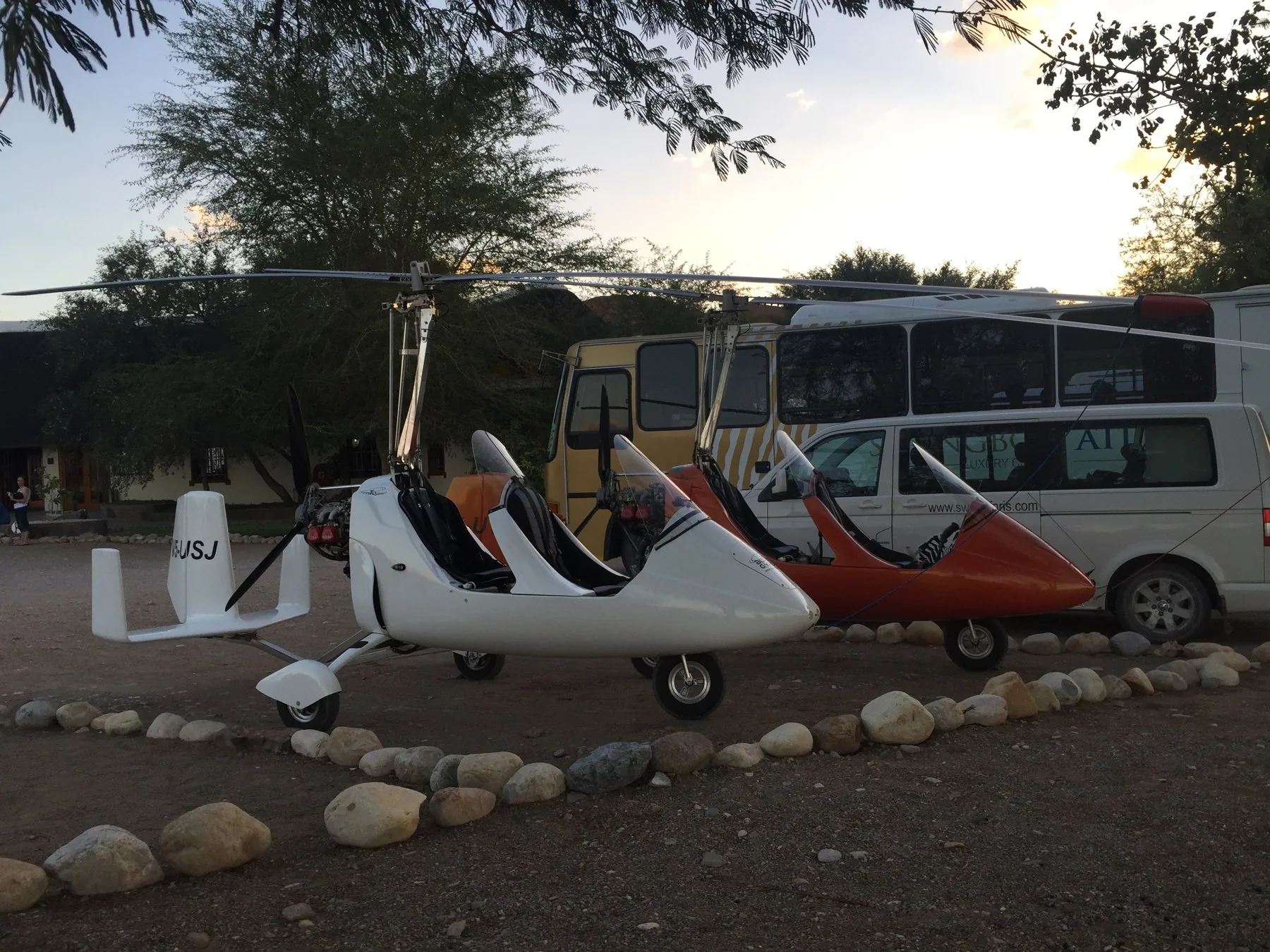 Gyroplanes parked beside a bus and vehicles in the shade at a Namibian lodge